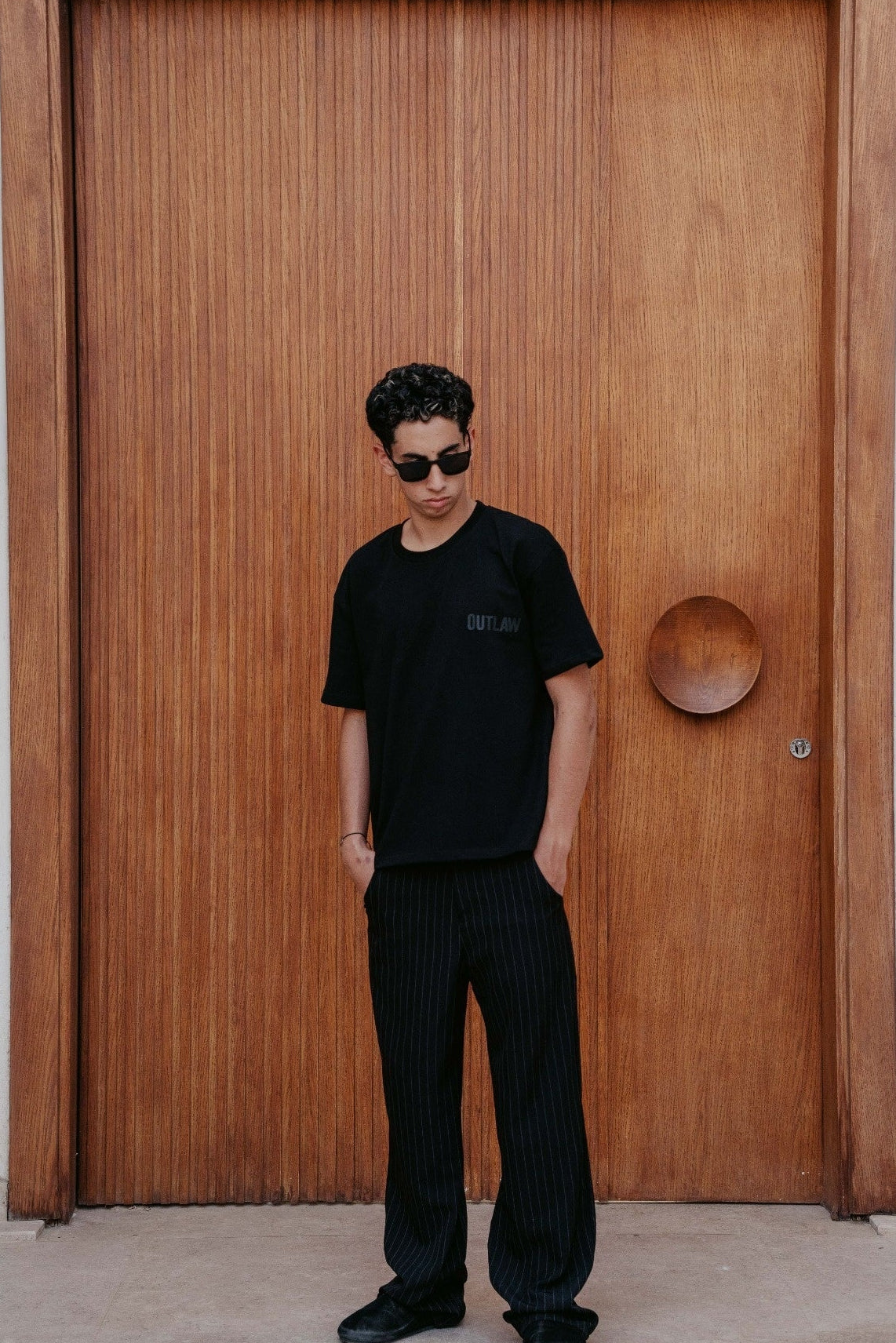 A man wearing a black short-sleeve t-shirt with a small chest logo and black pinstriped trousers, standing in front of a large wooden door.